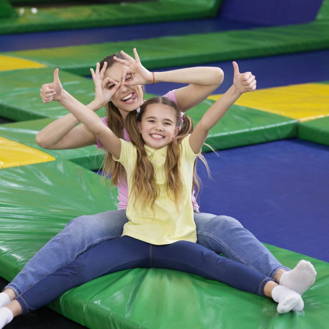 Playful silly mom with her teen daughter sitting at trampoline area of entertainment centre, showing